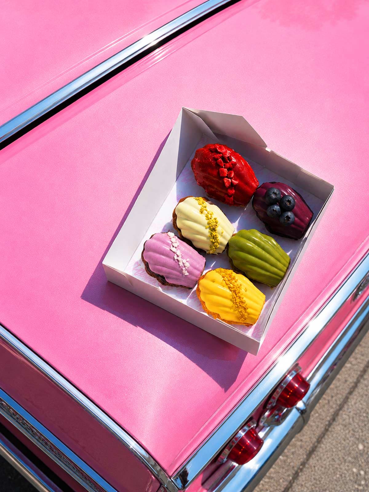 Box of colorful glazed madeleines on pink car hood